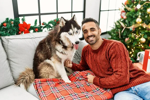 Young hispanic man smiling confident sitting on sofa with dog by christmas tree at home