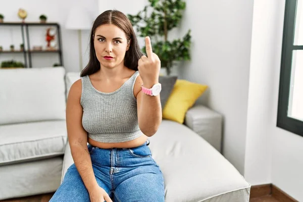 Young brunette woman sitting on the sofa at home showing middle finger, impolite and rude fuck off expression 