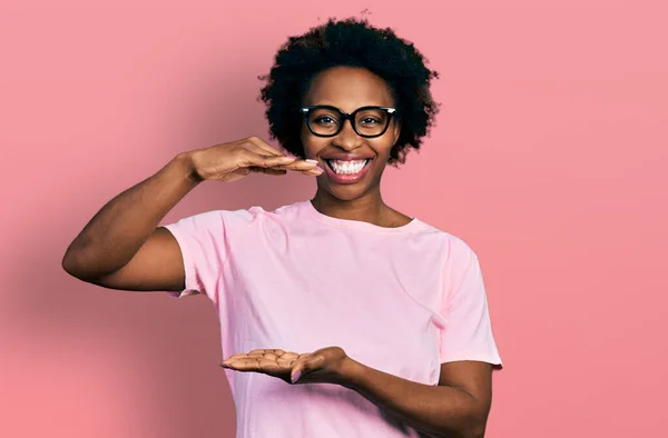 African american woman with afro hair wearing casual clothes and glasses gesturing with hands showing big and large size sign, measure symbol. smiling looking at the camera. measuring concept.