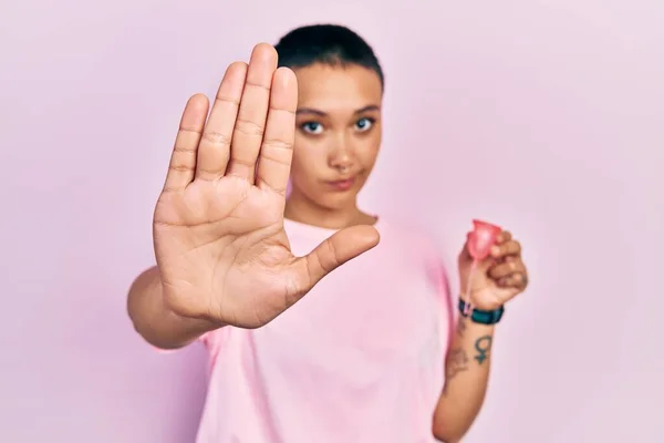 Beautiful hispanic woman with short hair holding menstrual cup with open hand doing stop sign with serious and confident expression, defense gesture