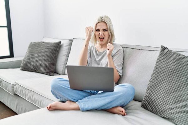 Young caucasian woman using laptop at home sitting on the sofa angry and mad raising fist frustrated and furious while shouting with anger. rage and aggressive concept. 