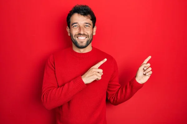 ハンサムな男で髭を生やしてカジュアルな赤いセーター笑顔とカメラを見て2本の手と指で側面を指して — ストック写真
