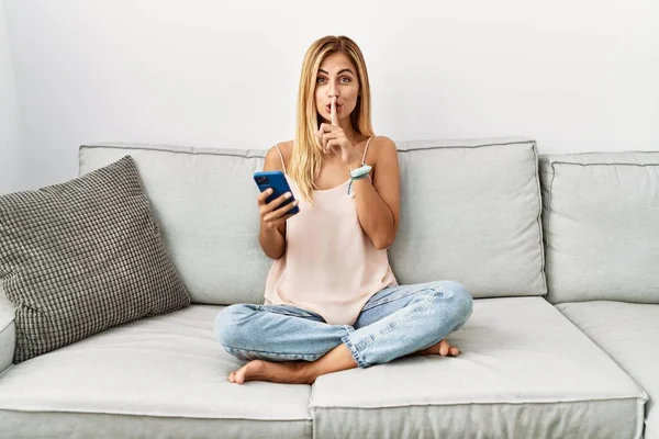 金发漂亮的年轻女人坐在家里的沙发上 用智能手机请求安静 手指放在嘴唇上 沉默和秘密概念 — 图库照片