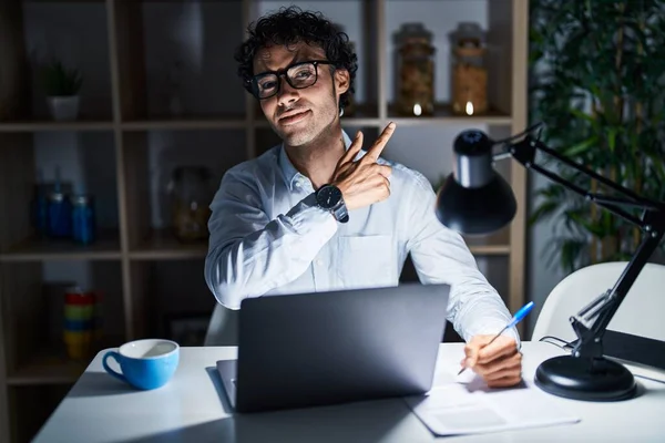 Hispanic man working at the office at night cheerful with a smile on face pointing with hand and finger up to the side with happy and natural expression