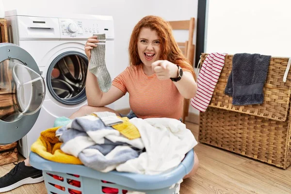若い赤毛の女性が洗濯機に汚れた洗濯機を入れて幸せと面白い顔でカメラに指を指しています 良いエネルギーと振動は — ストック写真