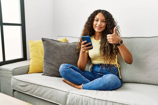 漂亮的惊慌失措的女人坐在家里的沙发上 用智能手机用手拿着快乐的大拇指做手势 赞成的表情看着相机显示的成功 — 图库照片