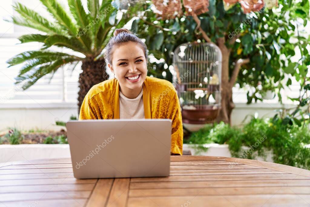 Beautiful young brunette woman using laptop sitting on the terrace at home looking positive and happy standing and smiling with a confident smile showing teeth 