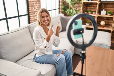 Young blonde woman recording vlog tutorial with smartphone at home smiling and looking at the camera pointing with two hands and fingers to the side. 
