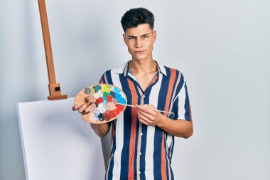 Ressam paleti ve boya fırçasını tutan genç İspanyol bir adam tuvale yakın duruyor şüpheci ve gergin, sorun yüzünden surat asıyor. negatif kişi. 