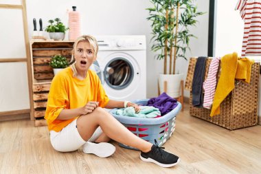 Young caucasian woman putting dirty laundry into washing machine in shock face, looking skeptical and sarcastic, surprised with open mouth 