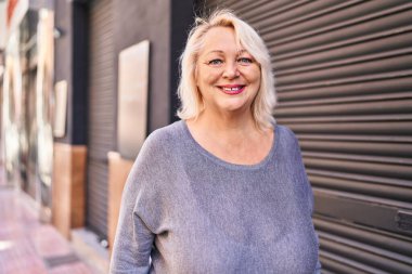 Orta yaşlı sarışın kadın sokakta kendinden emin bir şekilde gülümsüyor.