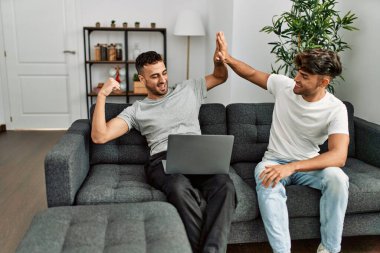 Two hispanic men couple using laptop high five with hands raised up at home