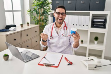 Young hispanic doctor man with beard holding ear cotton buds screaming proud, celebrating victory and success very excited with raised arms 