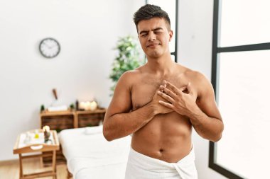 İspanyol asıllı genç adam spa merkezinde üstsüz duruyor. Elleri göğsünde, gözleri kapalı gülümsüyor ve yüzünde minnet ifadesi var. sağlık kavramı. 