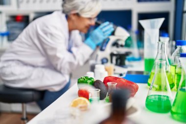 Laboratuvarda mikroskop kullanan orta yaşlı kadın bilim adamı üniforması giyiyor.