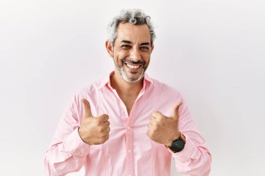 Orta yaşlı İspanyol bir adam arka planda izole edilmiş başarı işaretinin başında duruyor. Elleriyle olumlu jest yapıyor, gülümsüyor ve mutlu. Neşeli ifade ve kazanan jest. 