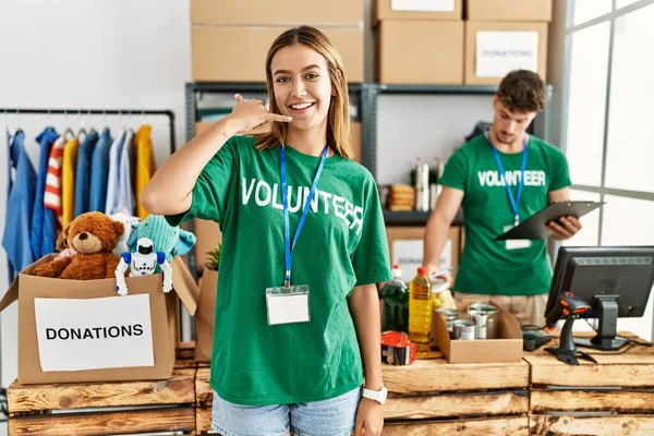 Young blonde girl wearing volunteer t shirt at donation stand smiling doing phone gesture with hand and fingers like talking on the telephone. communicating concepts.