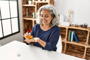 Middle age grey-haired woman taking pills sitting on the table at home.