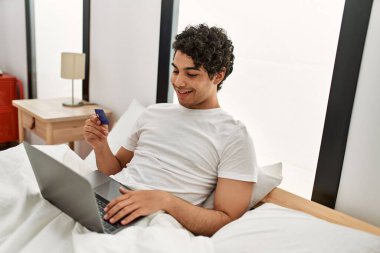 Laptop ve kredi kartı kullanan İspanyol bir adam yatak odasındaki yatakta oturuyor..