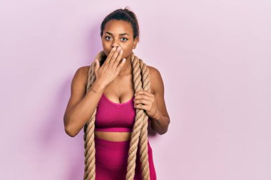Young african american girl training with battle rope covering mouth with hand, shocked and afraid for mistake. surprised expression 