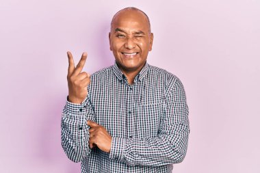 Orta yaşlı, sıradan kıyafetler giyen, gülümseyen, kameraya göz kırpan, zafer işareti yapan Latin bir adam. İki numara.. 