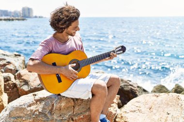 Genç İspanyol adam klasik gitar çalıyor plajda rock 'ın üzerinde oturuyor..