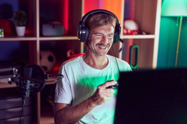 Genç sarışın adam oyun odasında joystick kullanarak video oyunu oynuyor.