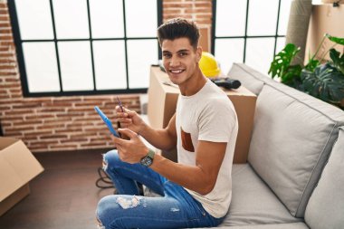 Young hispanic man using touchpad and credit card sitting on sofa at new home