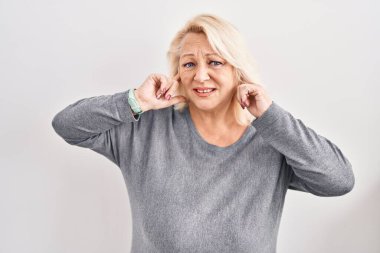 Middle age caucasian woman standing over white background covering ears with fingers with annoyed expression for the noise of loud music. deaf concept. 