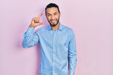 Hispanic man with beard wearing casual blue shirt smiling and confident gesturing with hand doing small size sign with fingers looking and the camera. measure concept. 
