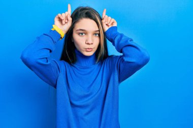Young brunette girl wearing turtleneck sweater doing funny gesture with finger over head as bull horns 