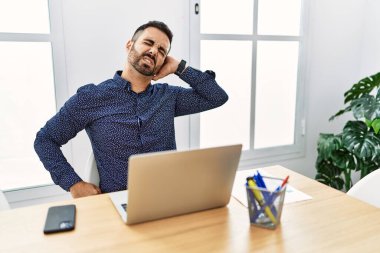 Young hispanic man with beard working at the office with laptop suffering of neck ache injury, touching neck with hand, muscular pain 