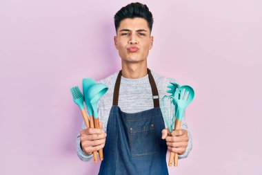 Young hispanic man wearing professional baker apron holding cooking tools looking at the camera blowing a kiss being lovely and sexy. love expression. 