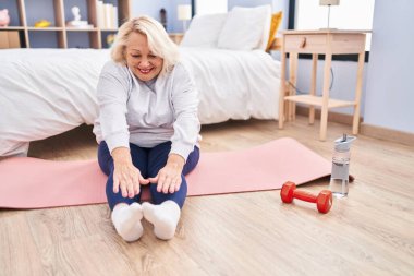 Orta yaşlı sarışın kadın gülümsüyor yatak odasında esniyor.