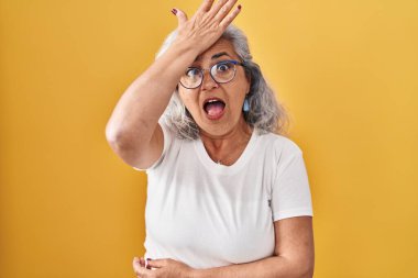 Middle age woman with grey hair standing over yellow background surprised with hand on head for mistake, remember error. forgot, bad memory concept. 