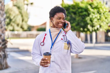 Afro-Amerikan kadın doktor üniforması giyiyor akıllı telefonla konuşuyor parkta kahve içiyor.