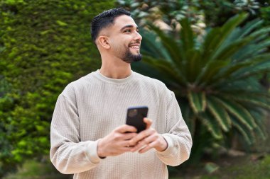 Genç Arap adam parkta akıllı telefon kullanarak kendinden emin gülümsüyor.