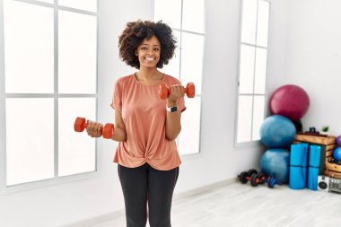 Genç Afrikalı Amerikalı kadın gülümsüyor spor merkezinde halterleri kullanarak kendine güven eğitimi alıyor