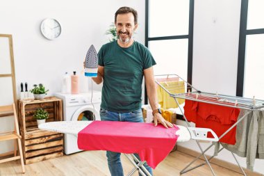 Middle age man with beard ironing clothes at home looking positive and happy standing and smiling with a confident smile showing teeth 