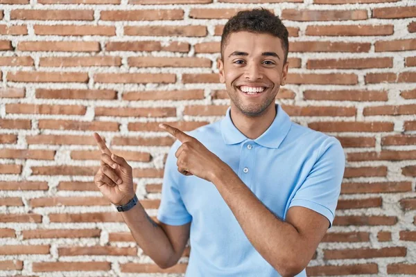 ブラジルの若者がレンガの壁の上に立って笑ってカメラを見て両手と指を横に向けて — ストック写真