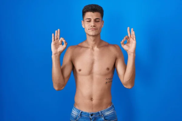 Young hispanic man standing shirtless over blue background relaxed and smiling with eyes closed doing meditation gesture with fingers. yoga concept.