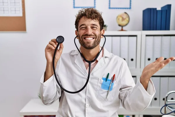 Young hispanic doctor man wearing doctor uniform holding stethoscope at clinic celebrating achievement with happy smile and winner expression with raised hand