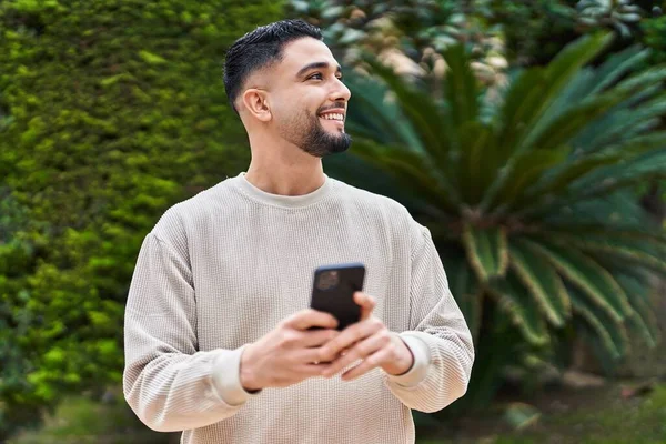 Genç Arap adam parkta akıllı telefon kullanarak kendinden emin gülümsüyor.