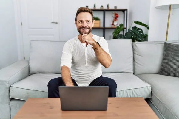 Middle age man using laptop at home with hand on chin thinking about question, pensive expression. smiling and thoughtful face. doubt concept.