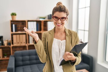 Young woman working at consultation office smiling cheerful presenting and pointing with palm of hand looking at the camera. 