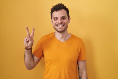 Sarı arka planda dikilen genç İspanyol adam iki numarayı gösterip işaret ederken kendinden emin ve mutlu bir şekilde gülümsüyor.. 