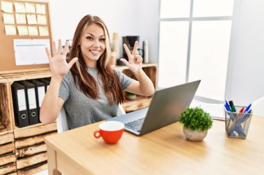 Young brunette woman working at the office with laptop showing and pointing up with fingers number eight while smiling confident and happy. 