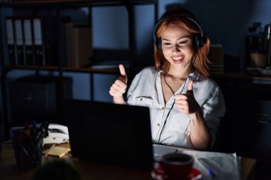 Young caucasian woman working at the office at night approving doing positive gesture with hand, thumbs up smiling and happy for success. winner gesture. 