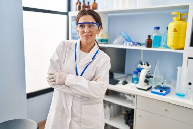 Laboratuvarda kollarını kavuşturmuş duran bilim adamı üniforması giyen İspanyol genç bir kadın.