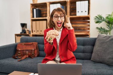 Young brunette woman at consultation office holding brain surprised with an idea or question pointing finger with happy face, number one 